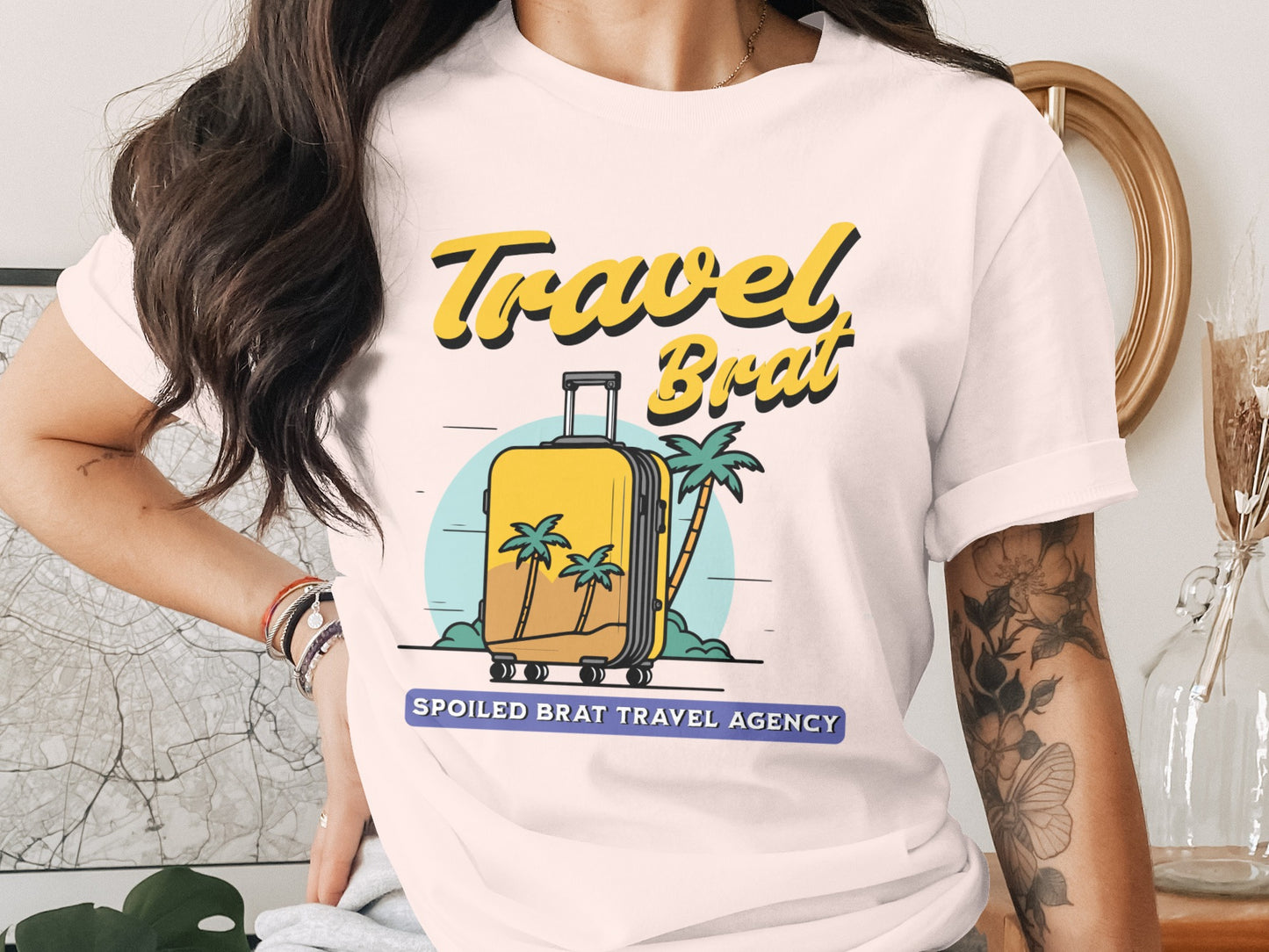 White t-shirt graphic with yellow text "Travel Brat" and a yellow suitcase on a beach.