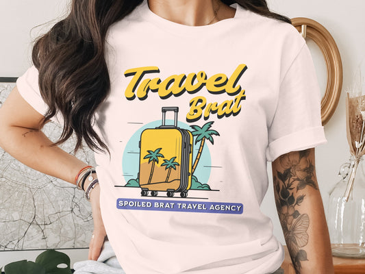 White t-shirt graphic with yellow text "Travel Brat" and a yellow suitcase on a beach.