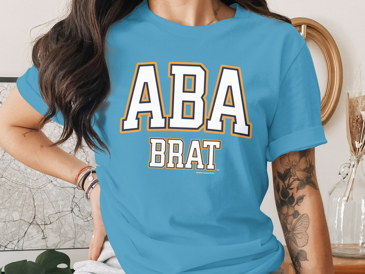 Light blue t-shirt with "ABA BRAT" in white, navy, and yellow lettering.