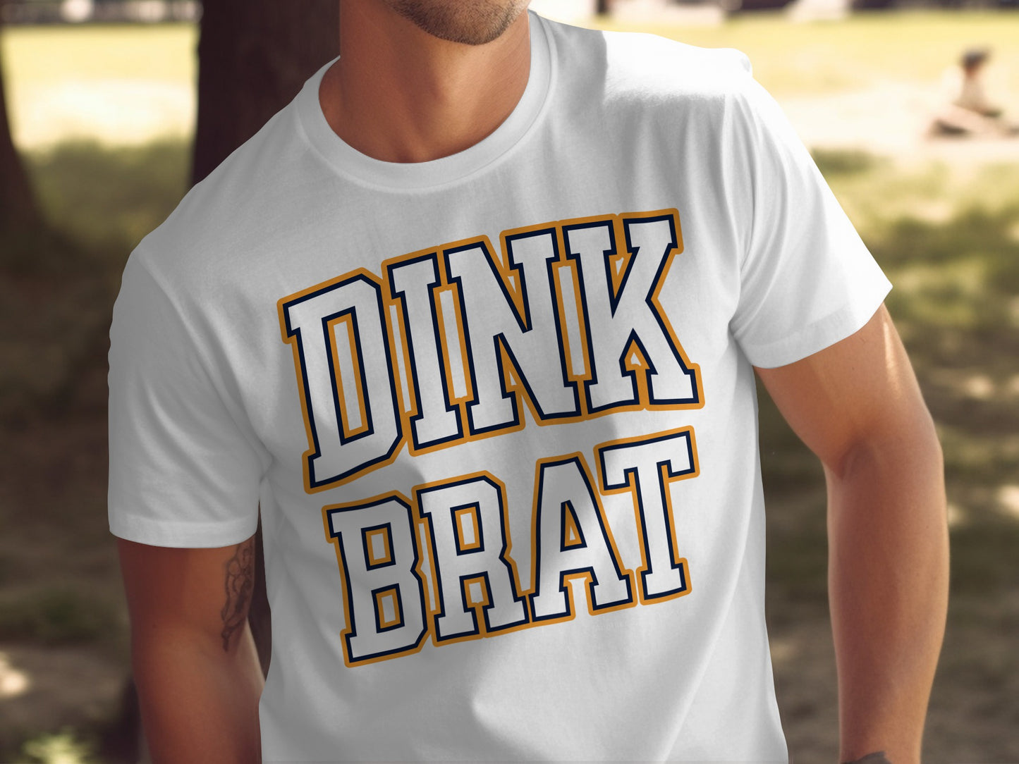 White t-shirt with navy and gold text that reads "DINK BRAT".
