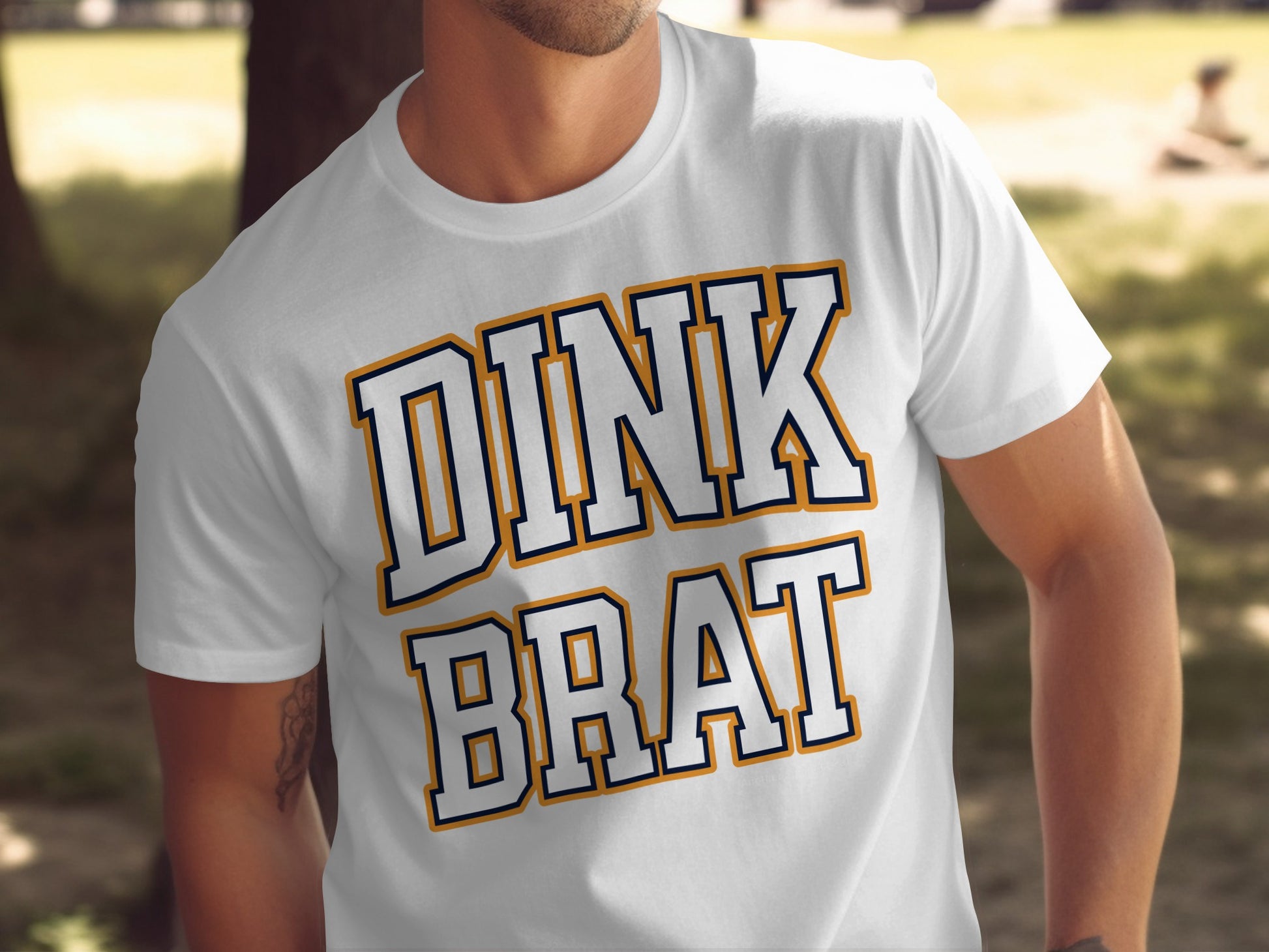 White t-shirt with navy and gold text that reads "DINK BRAT".
