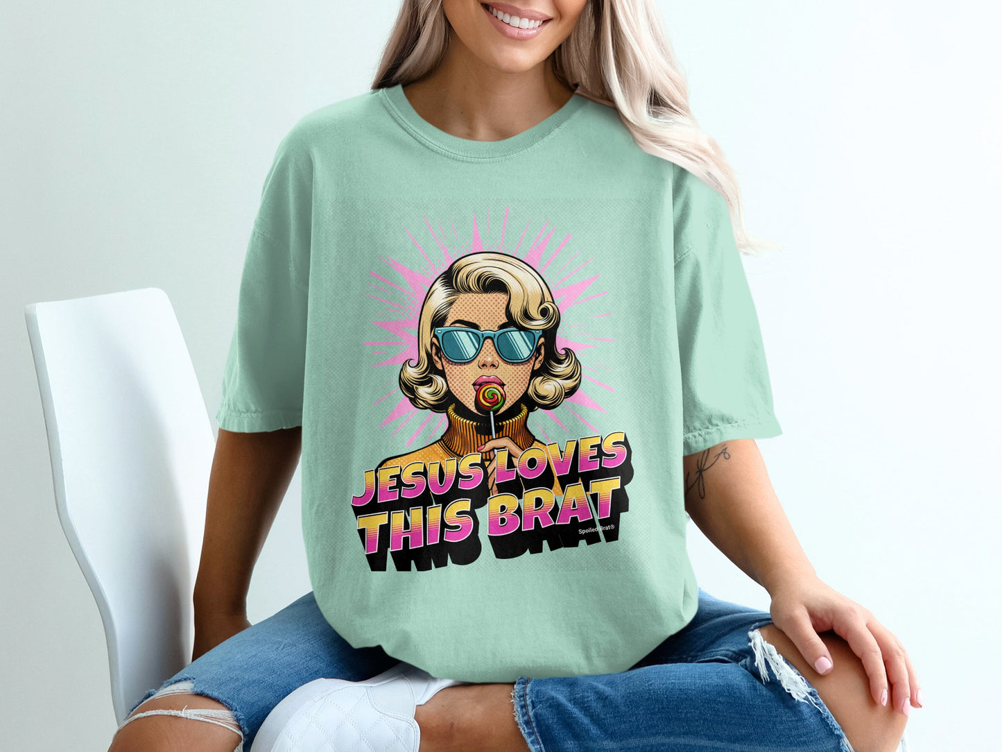 Mint t-shirt with retro pop art graphic of a woman holding a lollipop and text "JESUS LOVES THIS BRAT".