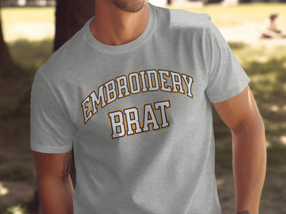Grey t-shirt with arched "EMBROIDERY" and "BRAT" text in white and orange outlines.