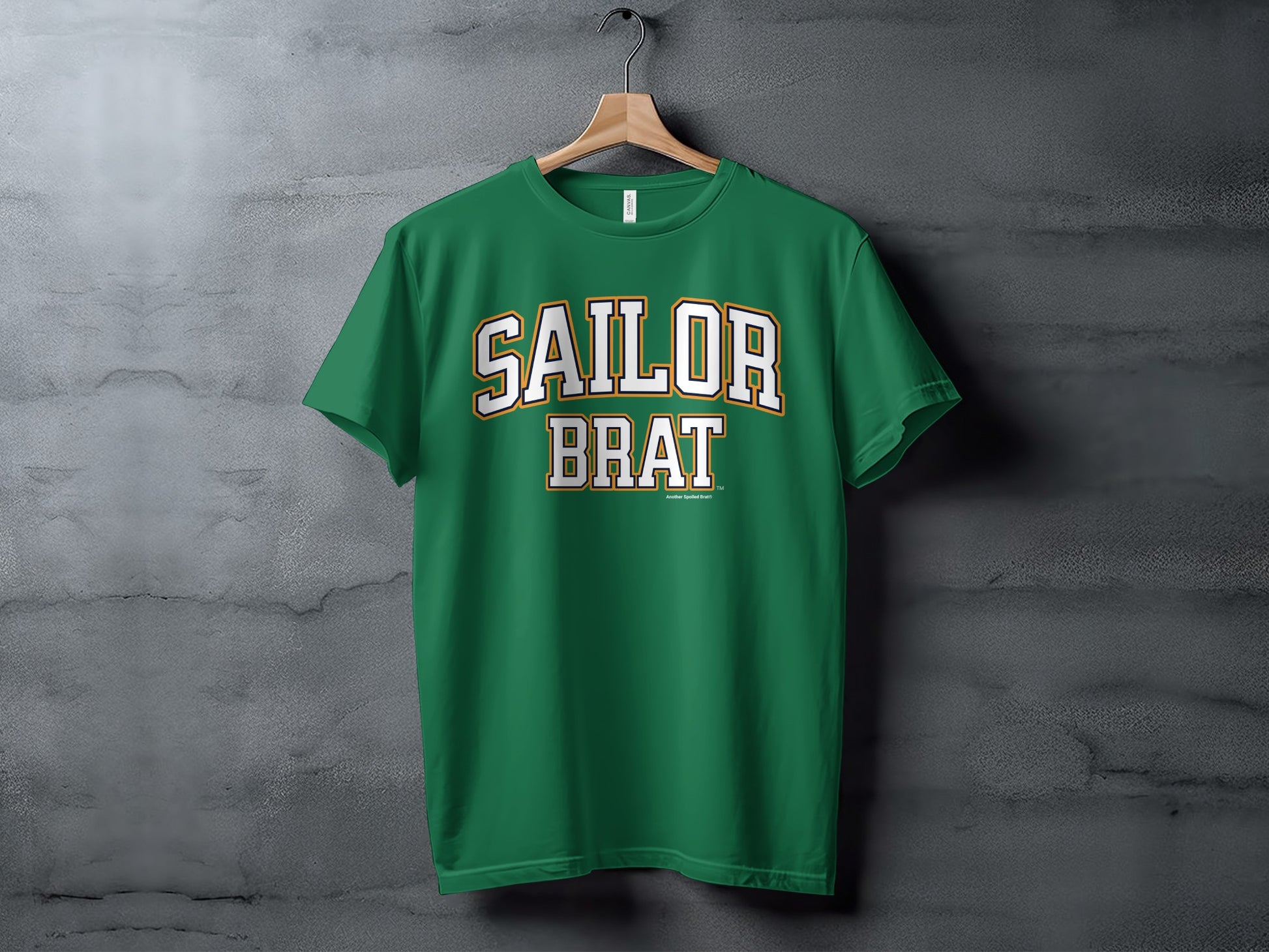 A green t-shirt with Sailor Brat printed in white and yellow block lettering hanging against a textured gray wall.