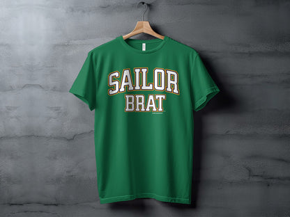 A green t-shirt with Sailor Brat printed in white and yellow block lettering hanging against a textured gray wall.
