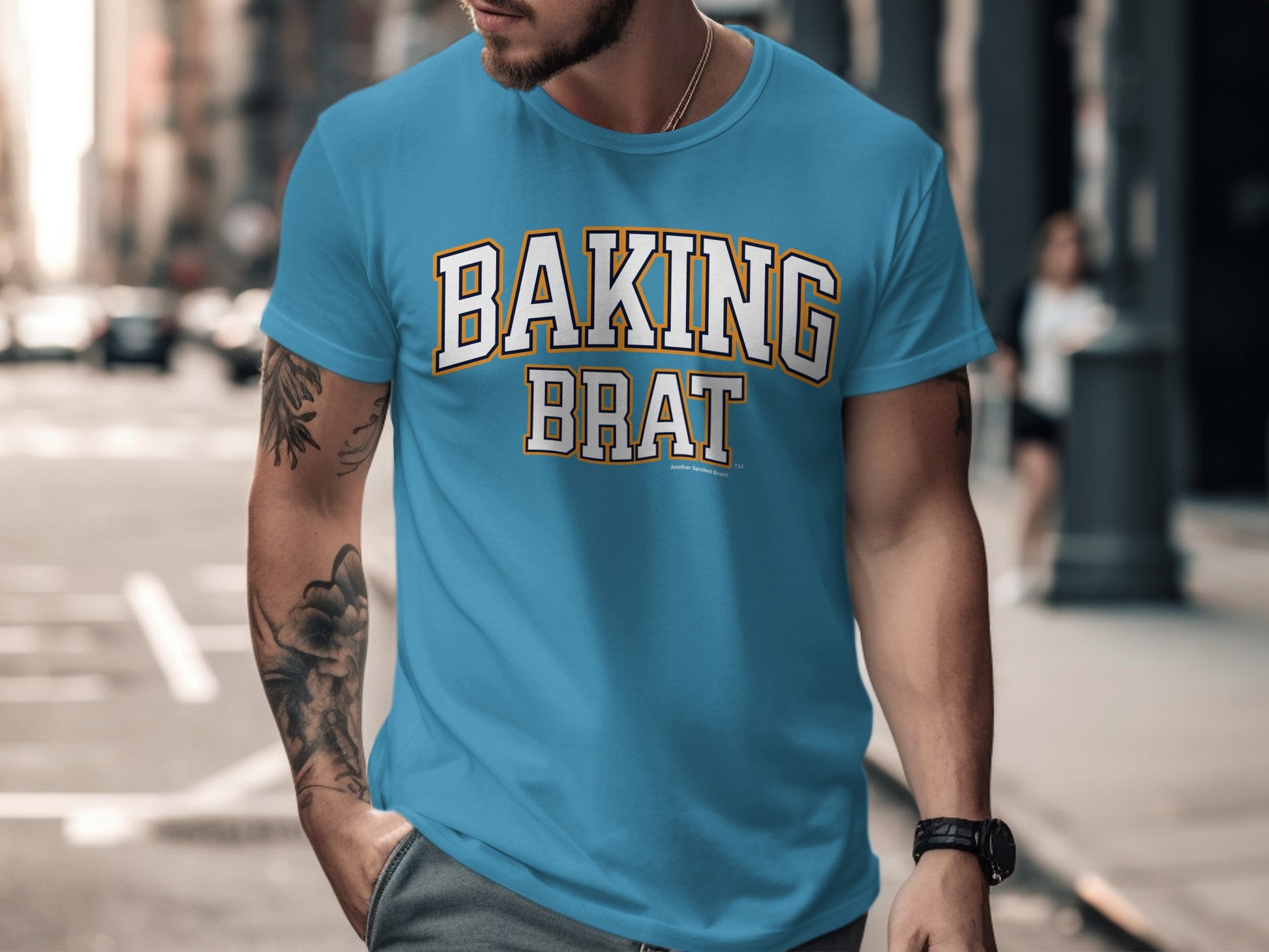 Turquoise t-shirt with "BAKING BRAT" text in white and orange on a blurred urban background.