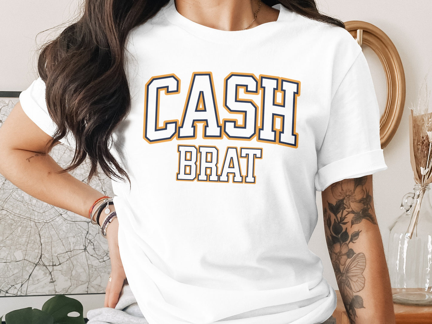 White t-shirt with navy and gold bold varsity text reading "CASH BRAT".