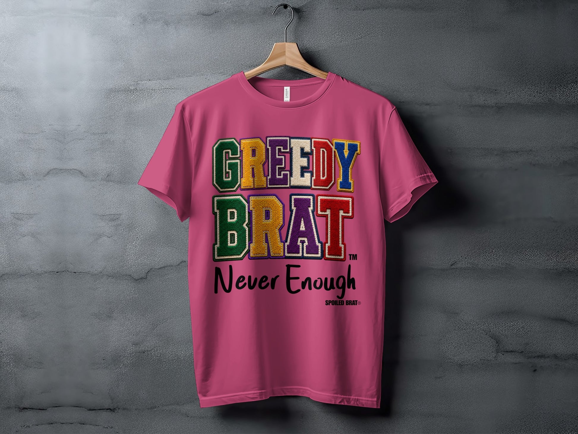 Pink t-shirt with "GREEDY BRAT Never Enough" text in colorful patch letters.