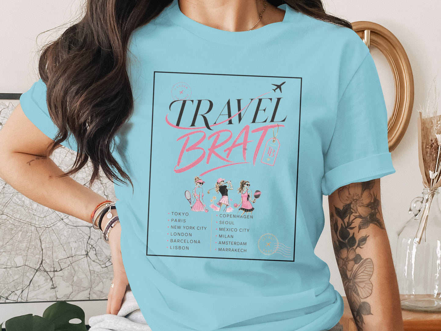 Light blue t-shirt with "TRAVEL BRAT" graphic and world cities, featuring three illustrated women.
