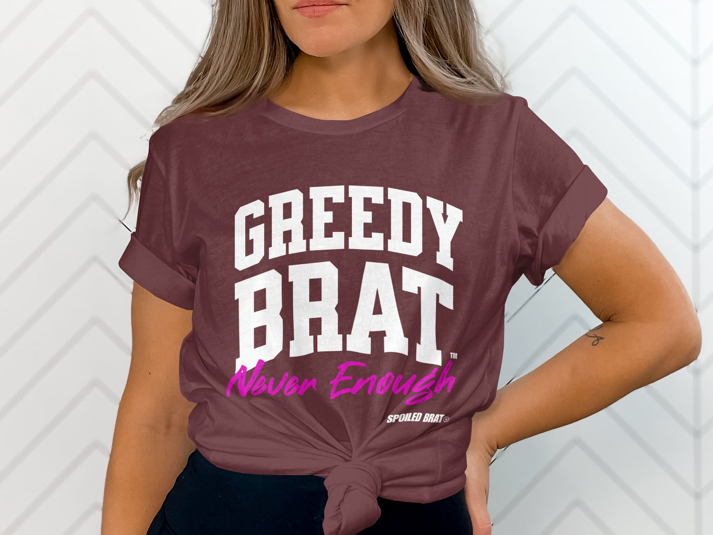 Maroon t-shirt with white and pink text reading "GREEDY BRAT Never Enough".