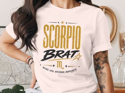 White t-shirt with "SCORPIO BRAT" and zodiac symbols in gold.