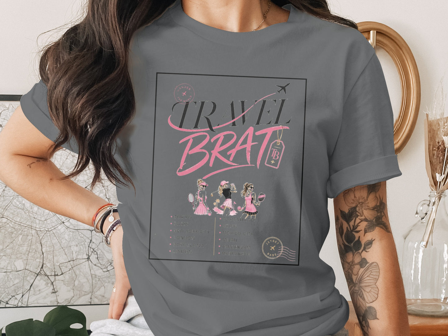 Grey t-shirt with "TRAVEL BRAT" graphic, three figures, and city list.