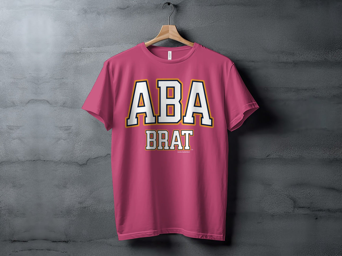 Pink t-shirt hanging on a wooden hanger, with "ABA BRAT" text.