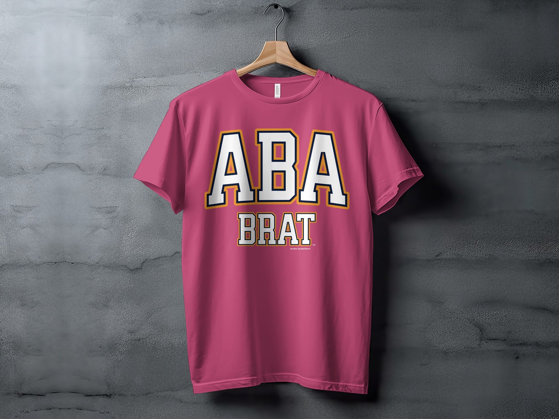 Pink t-shirt hanging on a wooden hanger, with "ABA BRAT" text.