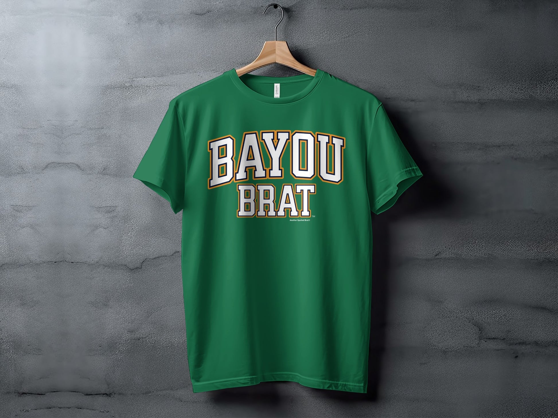 Green t-shirt with white and gold text reading "BAYOU BRAT" on a hanger.