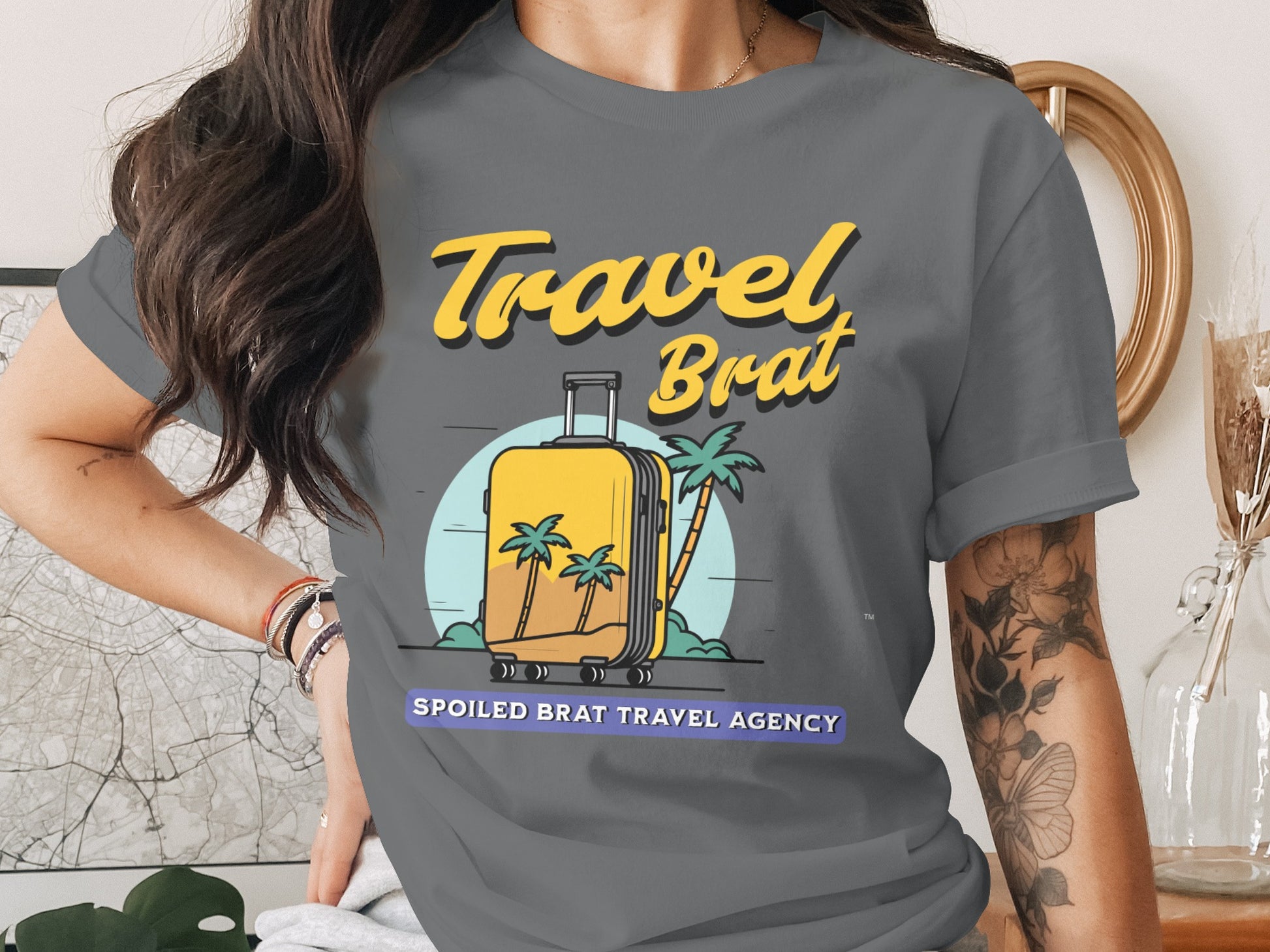 Grey t-shirt with yellow text "Travel Brat" and suitcase graphic on a tropical scene.