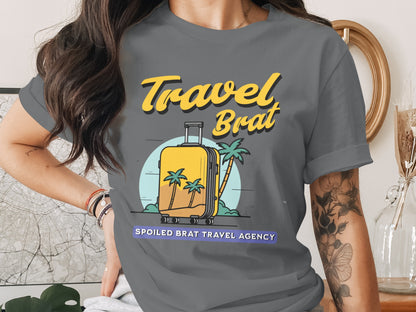 Grey t-shirt with yellow text "Travel Brat" and suitcase graphic on a tropical scene.