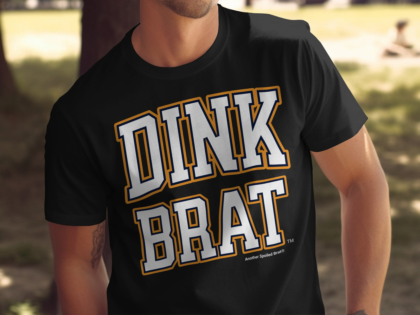 Black t-shirt with white and orange "DINK BRAT" text and "Another Spoiled Brat®" logo.
