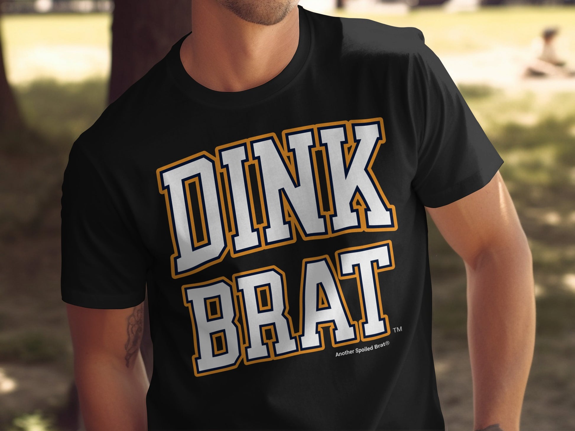 Black t-shirt with white and orange "DINK BRAT" text and "Another Spoiled Brat®" logo.