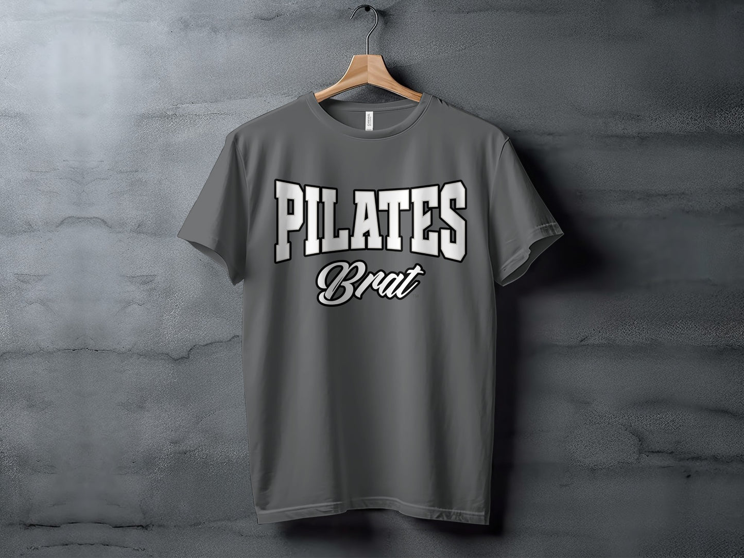 Gray t-shirt with "PILATES Brat" text, hanging on a wooden hanger against a textured wall.