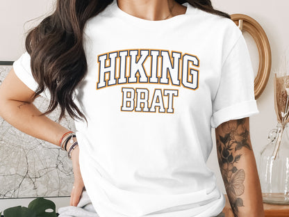 White t-shirt with "HIKING BRAT" text in navy and gold collegiate font.