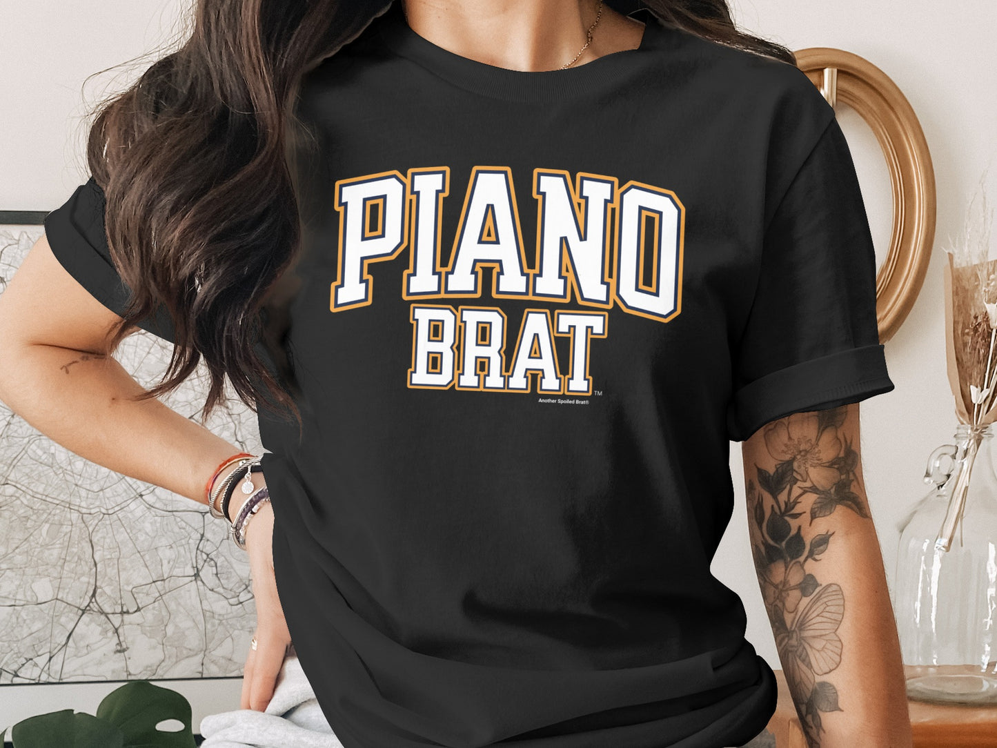 Black t-shirt with "PIANO BRAT" text in white and yellow collegiate font.