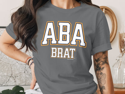 Grey t-shirt with white "ABA BRAT" text, outlined in yellow and navy.