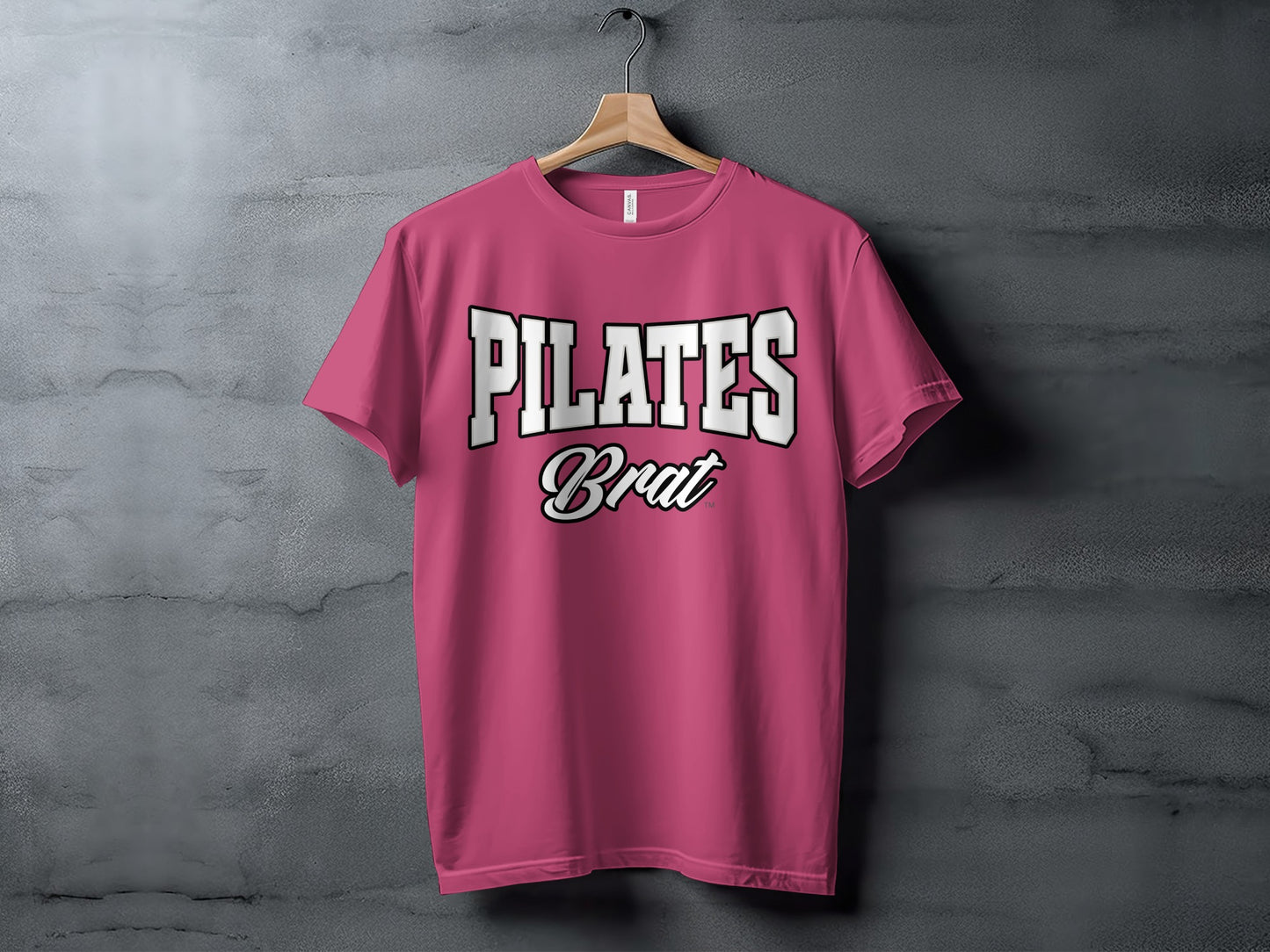 Pink t-shirt with white text that reads "PILATES Brat" displayed on a hanger.