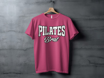 Pink t-shirt with white text that reads "PILATES Brat" displayed on a hanger.
