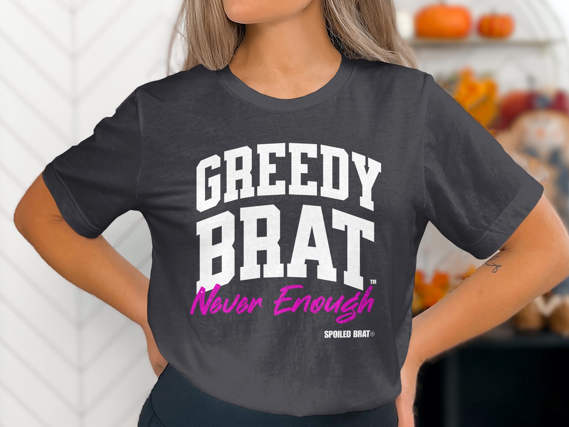 Dark grey t-shirt with white text "GREEDY BRAT" and pink text "Never Enough".