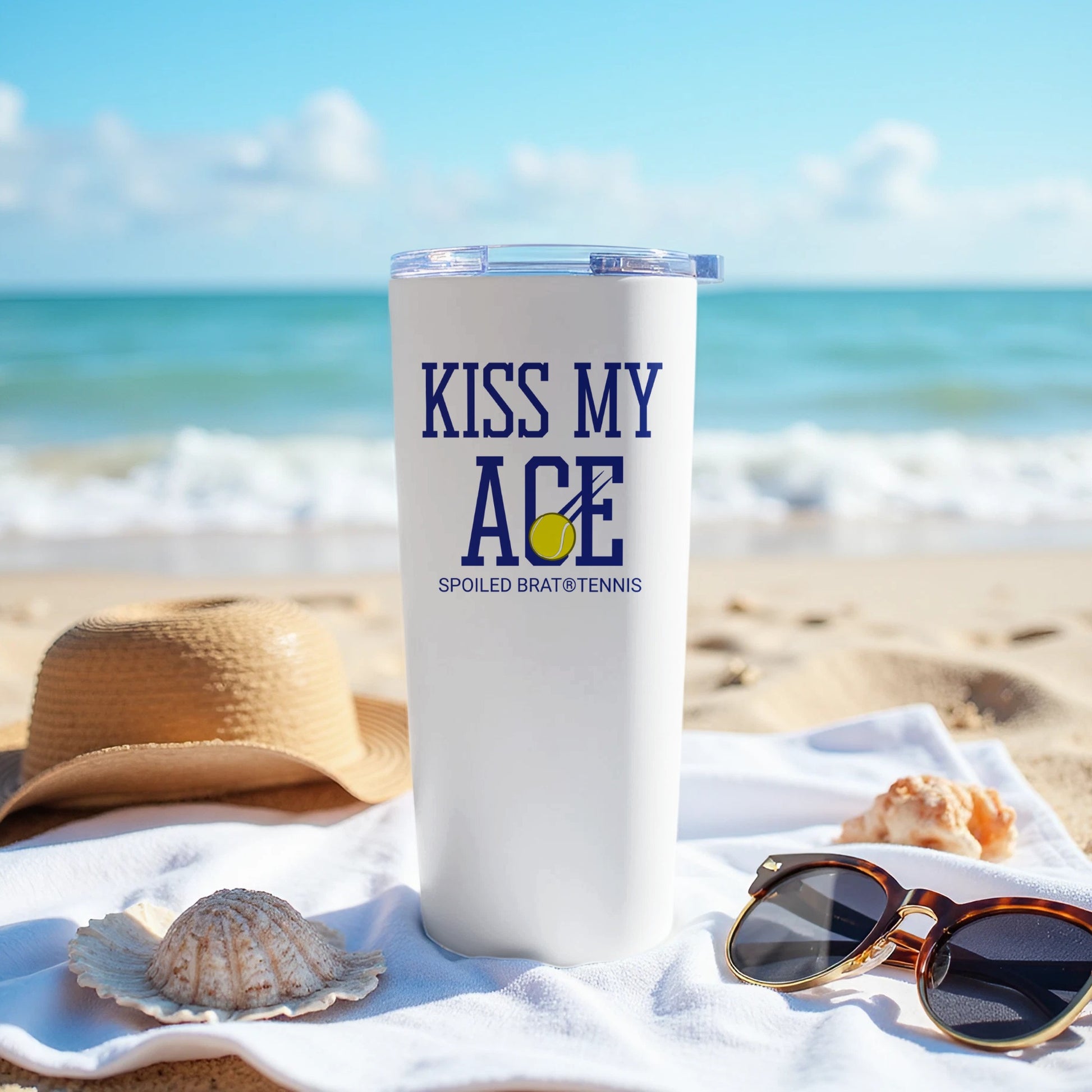 White tumbler with "KISS MY ACE" and tennis ball graphic on a beach.