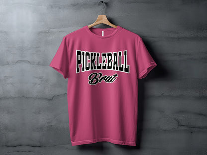 Pink t-shirt with "PICKLEBALL Brat" text design hanging on a hanger.