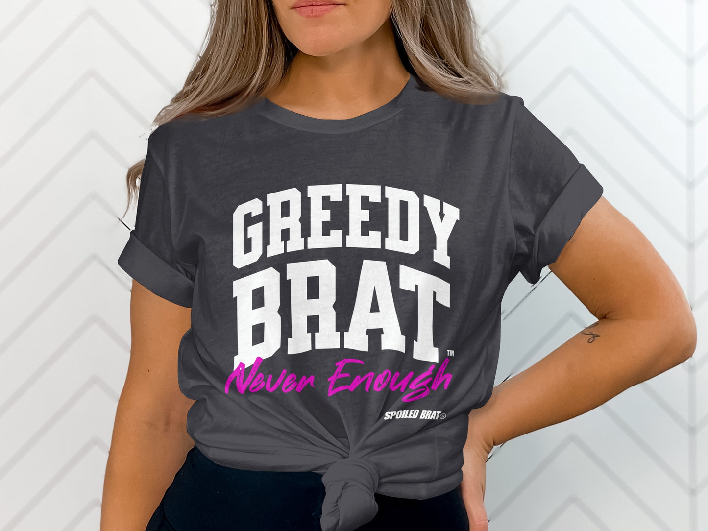 Dark grey t-shirt with white text "GREEDY BRAT" and pink text "Never Enough".
