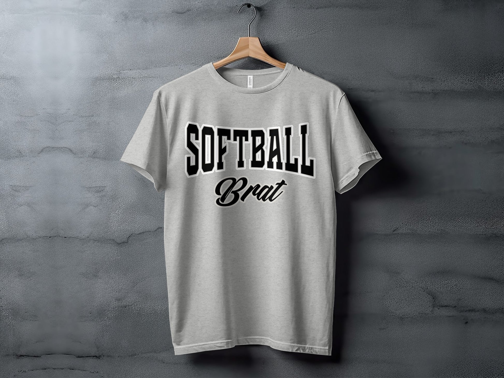 Light grey t-shirt with black text "SOFTBALL Brat" hanging on a wooden hanger against a concrete wall.