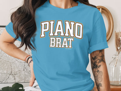 Light blue t-shirt with "PIANO BRAT" text in stylized white and orange letters.