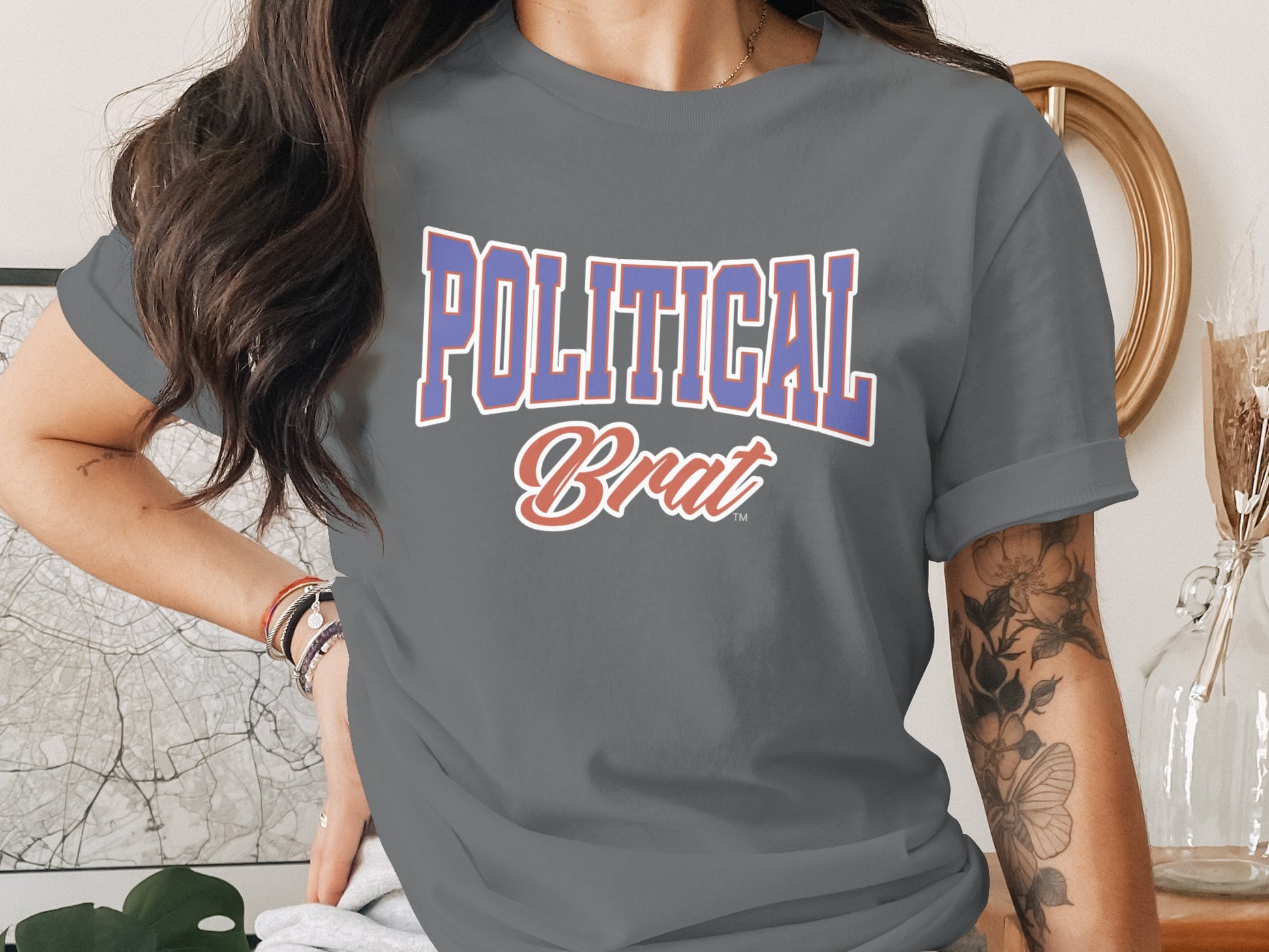 Graphic t-shirt with "POLITICAL Brat" text, worn by a person with tattoos and jewelry.