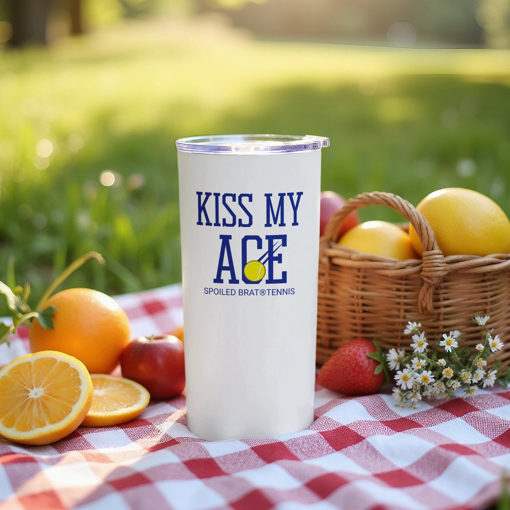 White tumbler with "Kiss My Ace" and tennis ball logo on picnic blanket.