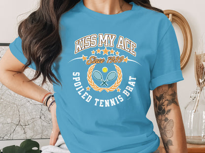 Blue t-shirt with "Kiss My Ace Spoiled Tennis Brat Since 1989" text and tennis graphics.