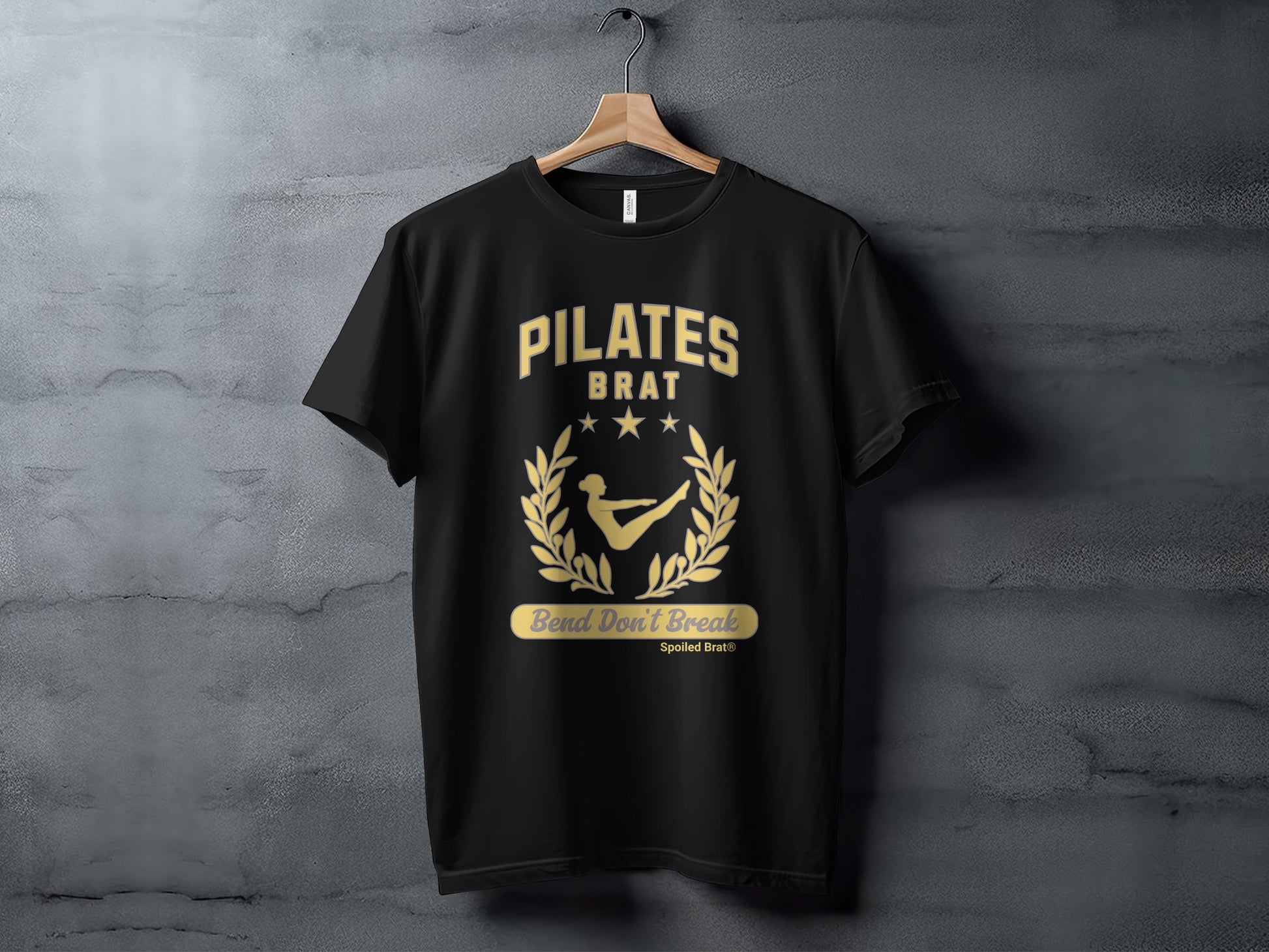 Black t-shirt with gold "PILATES BRAT" text and a laurel wreath with a figure.