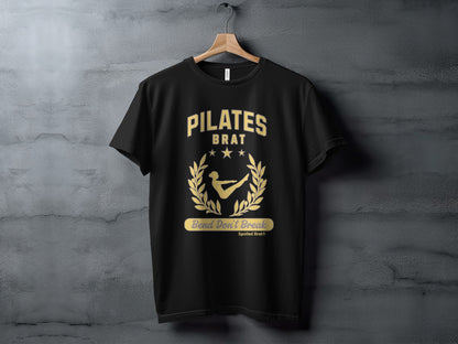 Black t-shirt with gold "PILATES BRAT" text and a laurel wreath with a figure.