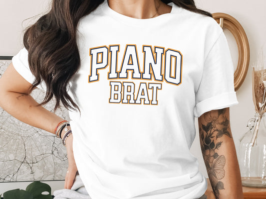 White t-shirt with "PIANO BRAT" text in collegiate font style.
