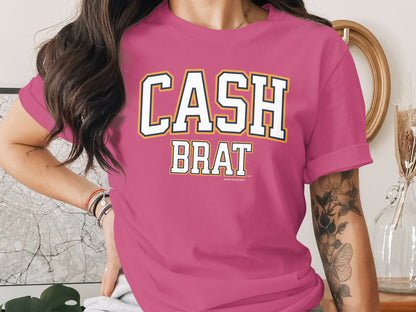 Pink t-shirt with "CASH BRAT" text in white, navy, and yellow block letters.