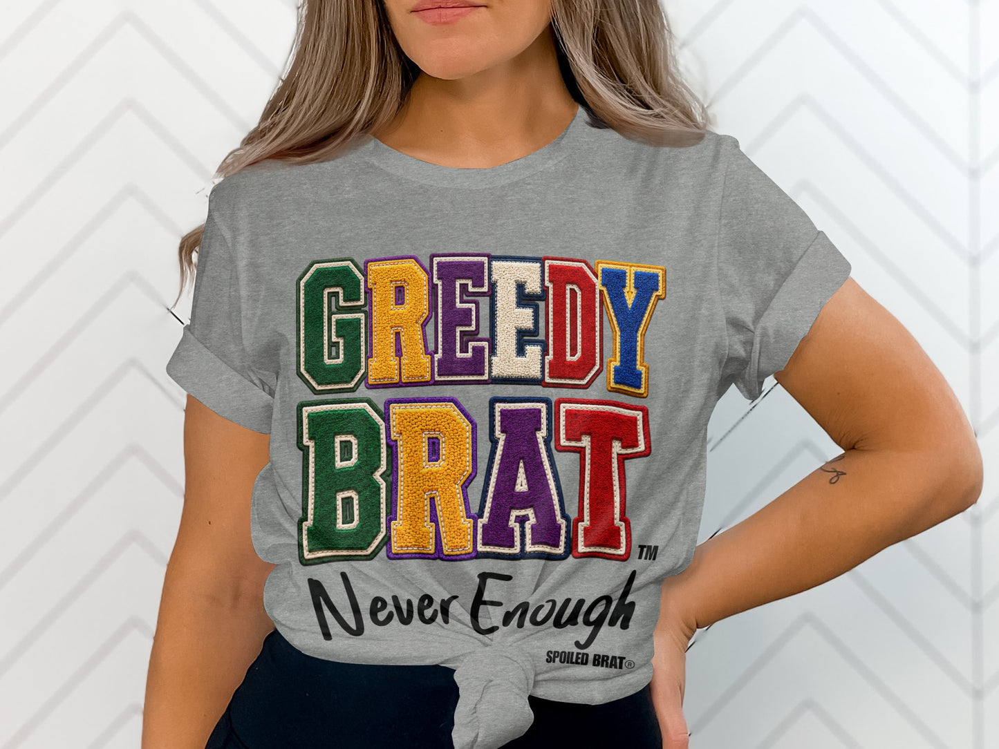 Grey t-shirt with "GREEDY BRAT Never Enough" text in colorful, appliquéd letters.