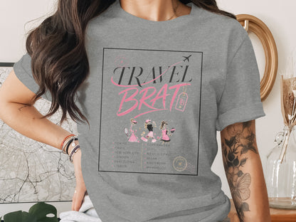 Grey t-shirt with "TRAVEL BRAT" graphic featuring city list and illustrated figures.
